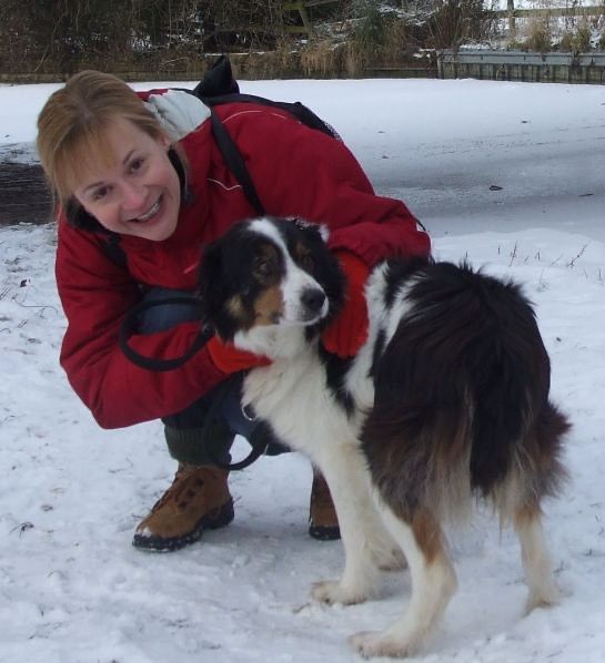 A person in a red jacket crouching in the snow while petting a tri-color dog on a leash.