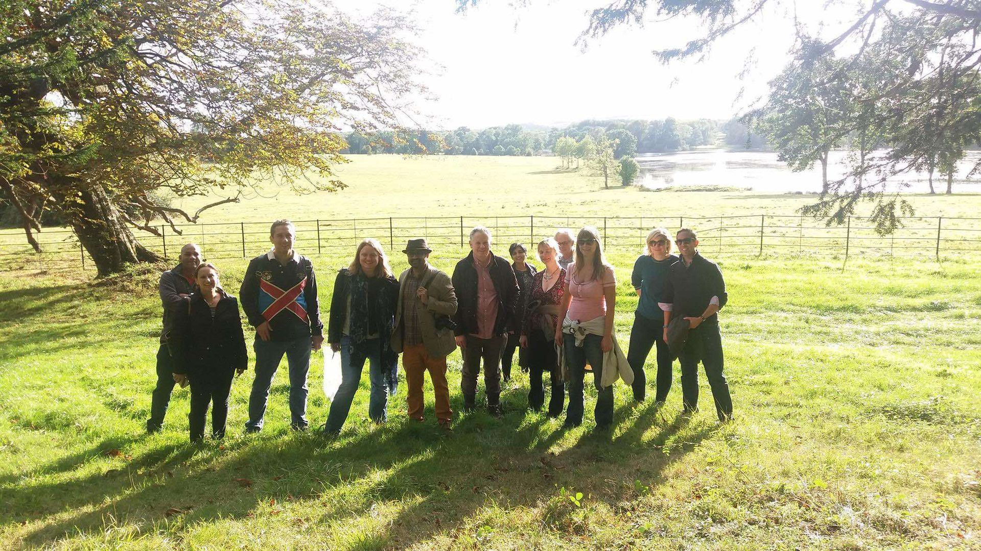 A group of twelve people standing in a sunny, grassy field with trees and a body of water in the background.