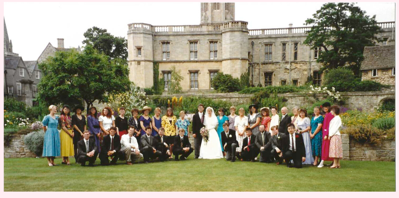 A large wedding party poses for a group photo on a grassy lawn in front of a stone mansion.