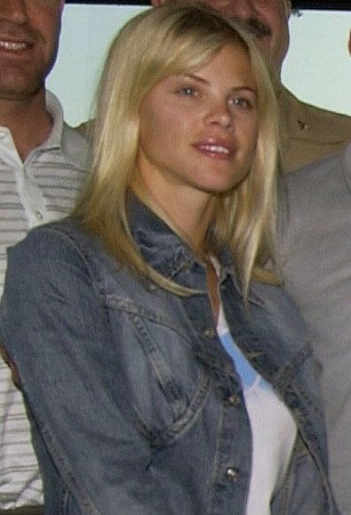 A person with blonde hair wearing a denim jacket and white shirt, smiling slightly while posing with others.