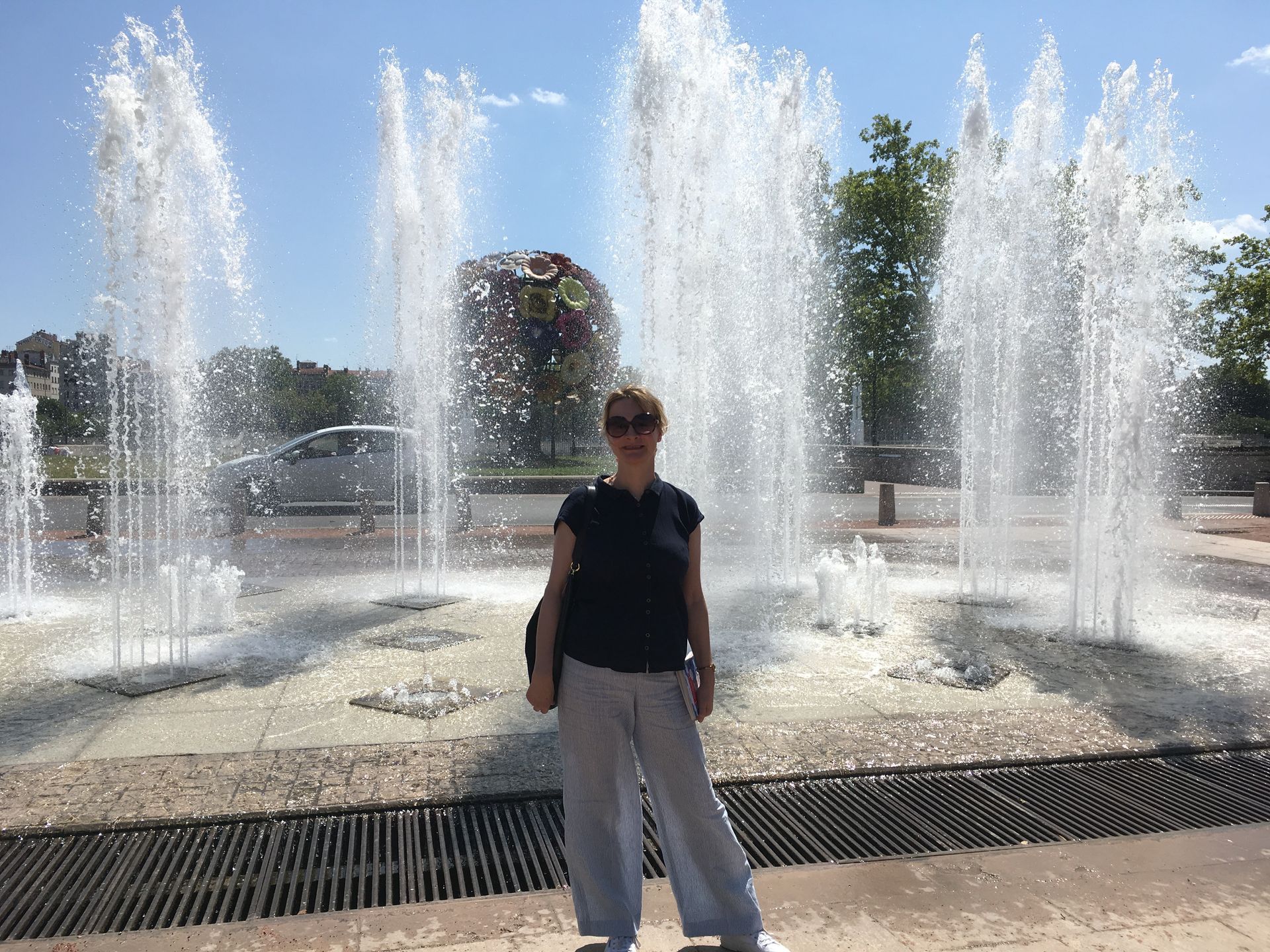 A person wearing sunglasses and a dark shirt stands in front of a series of tall, spraying water fountains on a sunny day.