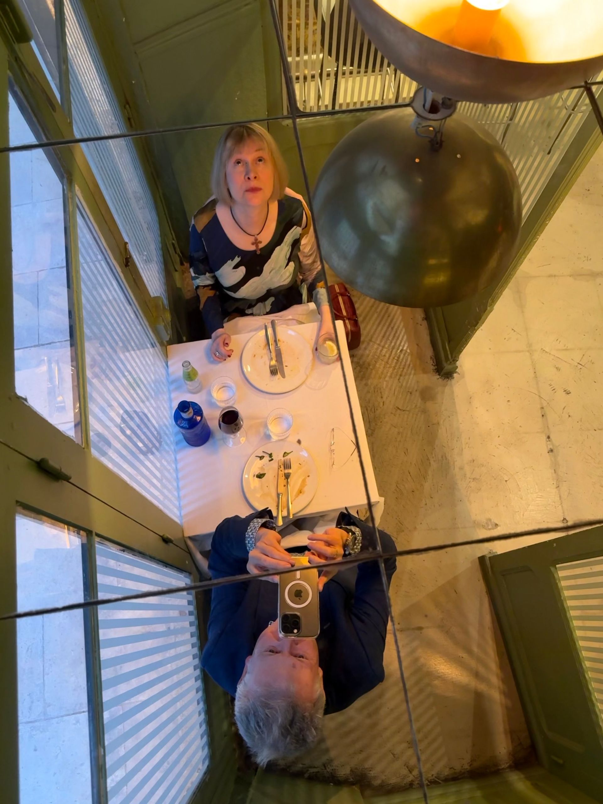 A high-angle view of two people at a restaurant table, one looking up and the other taking a photo with a smartphone.