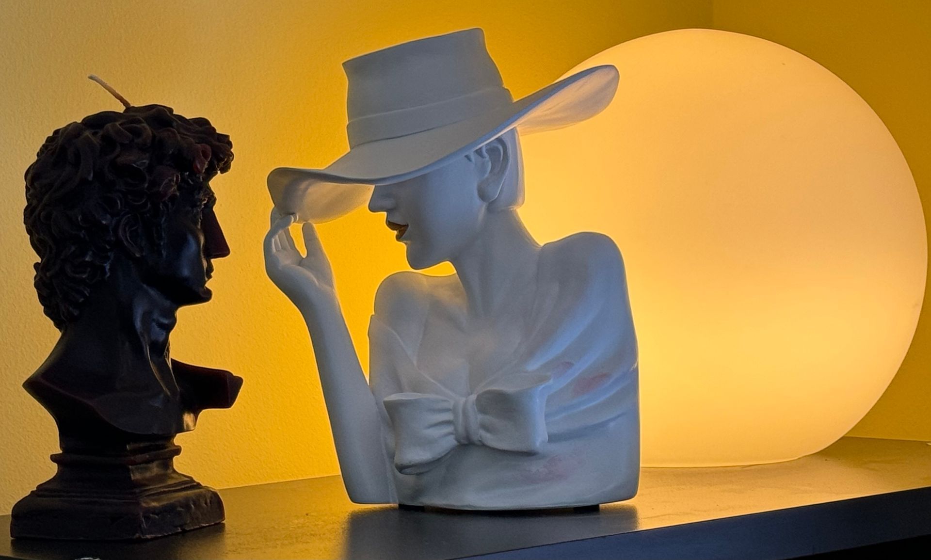 A black bust of a person next to a white sculpture of a figure in a wide-brimmed hat, set against a large glowing sphere.