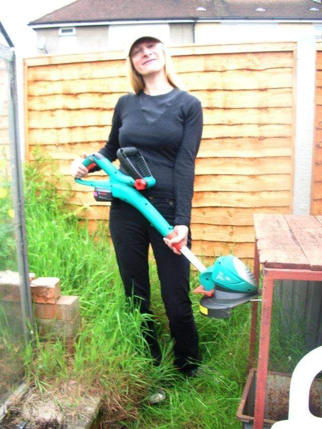 A person stands in a grassy garden holding a teal, battery-powered string trimmer, wearing a black top and pants and a cap.