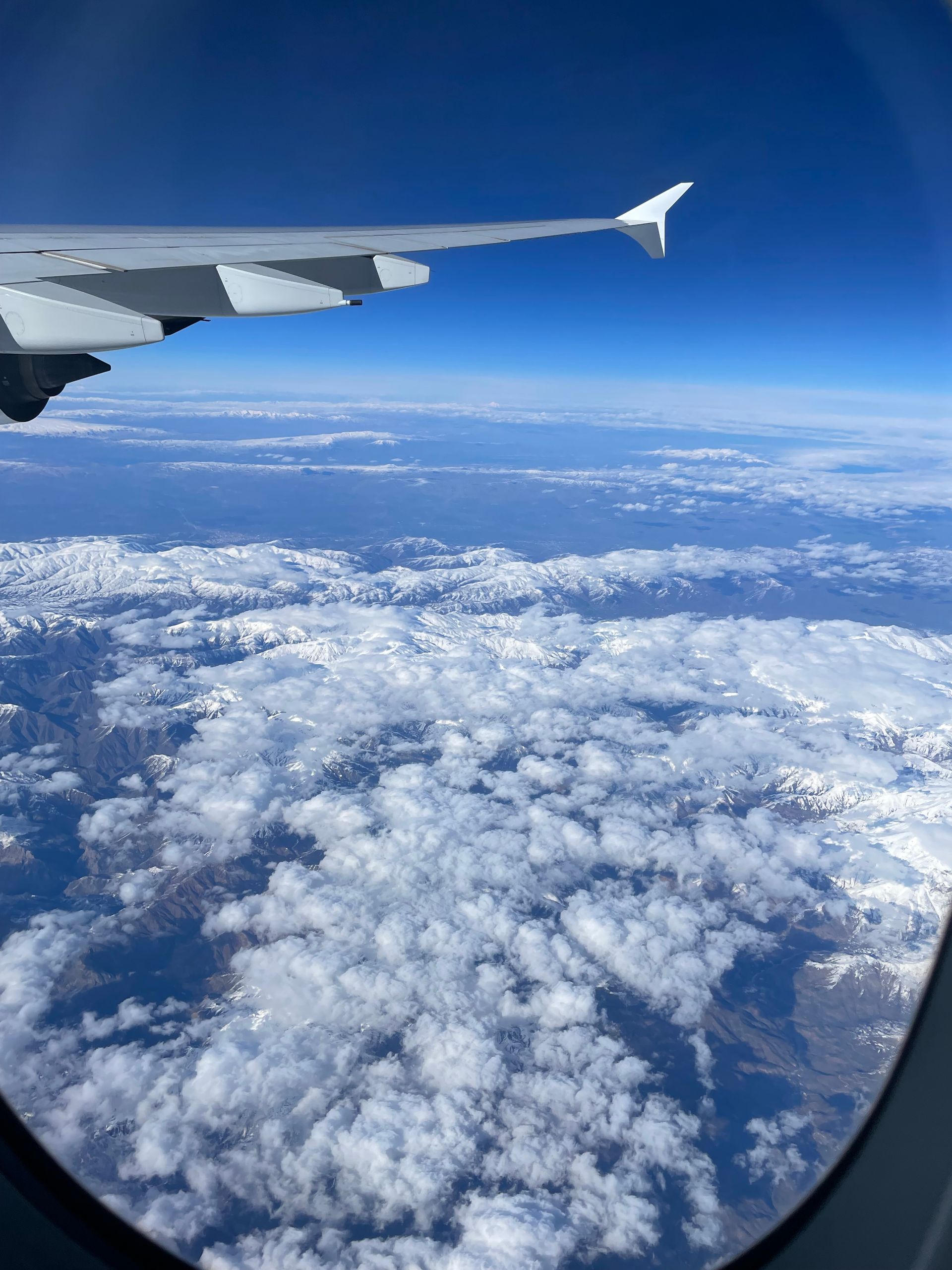 View from an airplane window showing a white wing over a vast landscape of snow-capped mountains and scattered clouds.