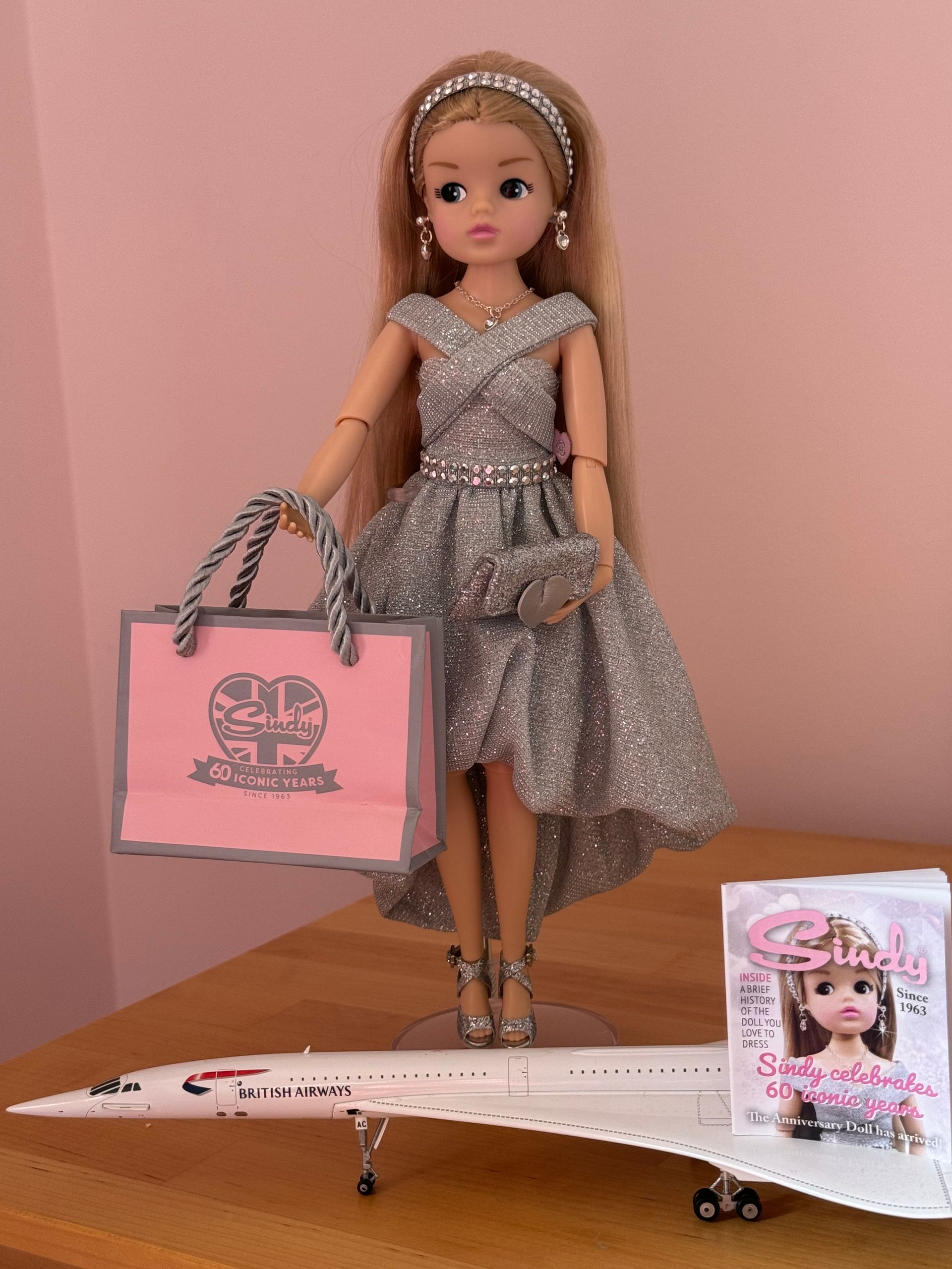 A blonde doll in a sparkling silver dress holds a pink shopping bag, standing beside a model Concorde plane.