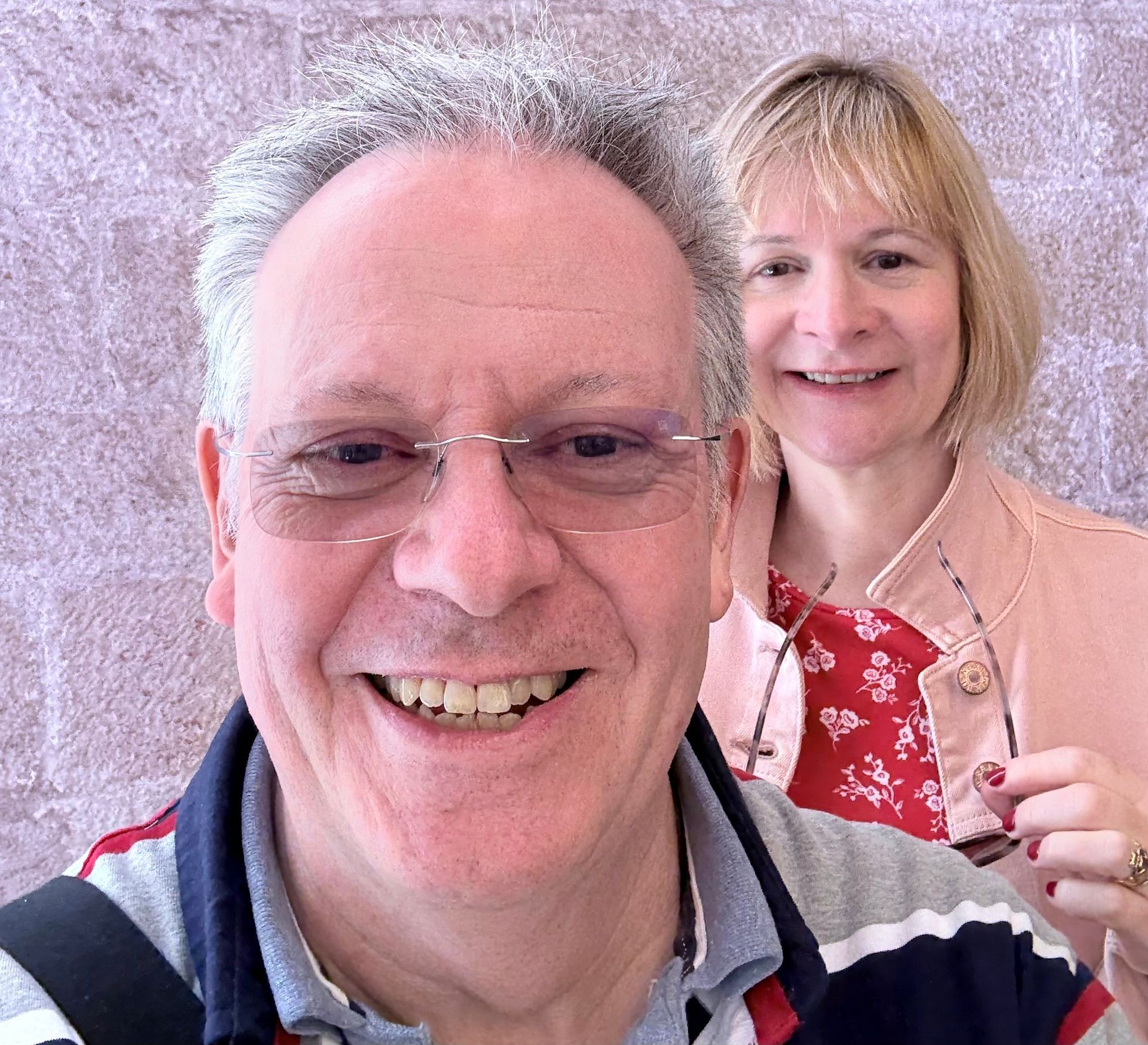 A man in a striped polo shirt smiles in the foreground, with a woman in a pink jacket standing behind him.