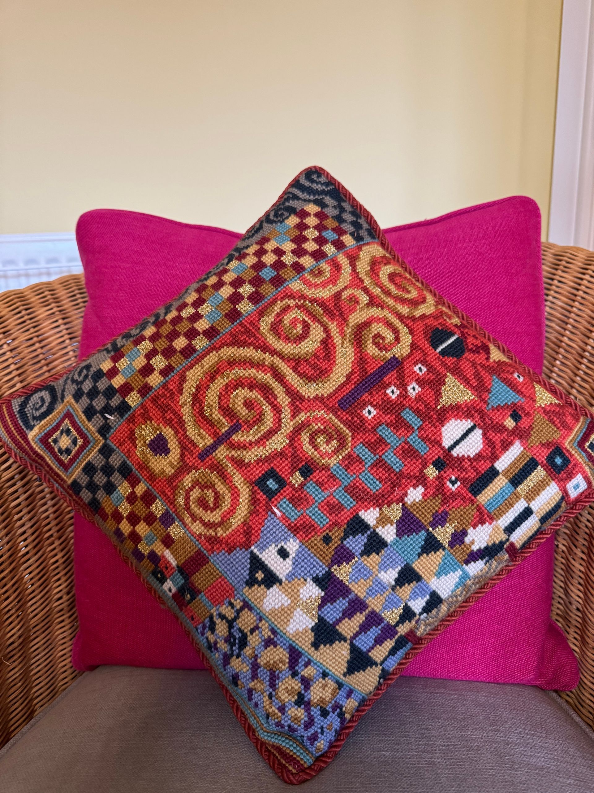 A square decorative cushion with ornate gold, red & blue geometric patterns resting in a wicker chair against a pink cushion.