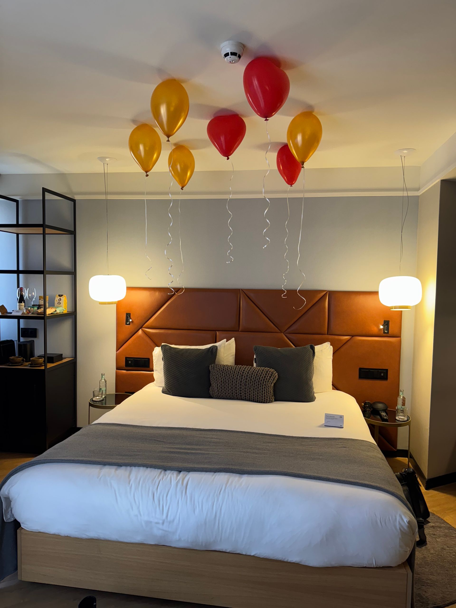 A hotel bed with a brown tufted headboard, decorated with red and yellow balloons floating above the pillows.
