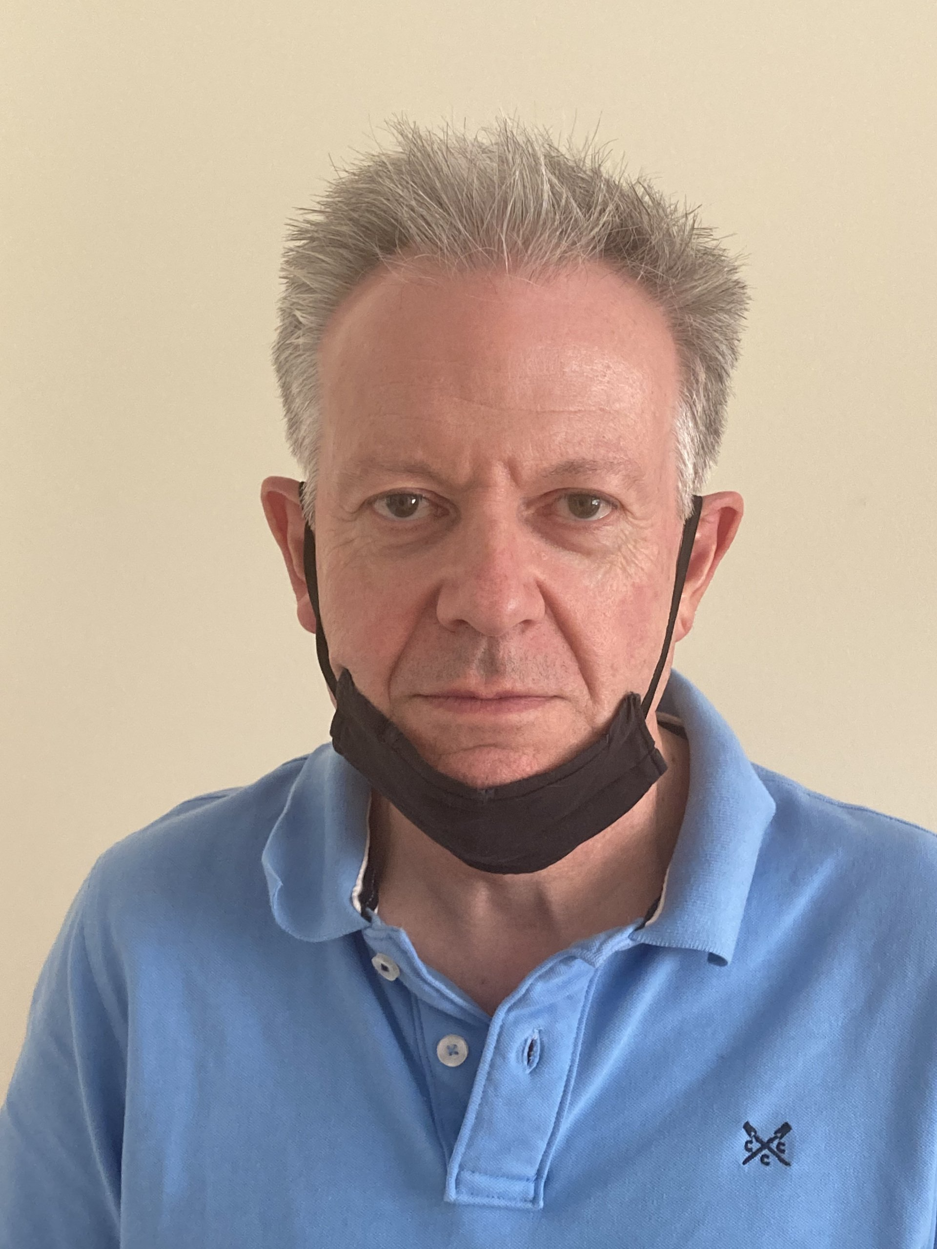 A person with short, light-colored hair wearing a light blue polo shirt and a black face mask pulled below their chin.