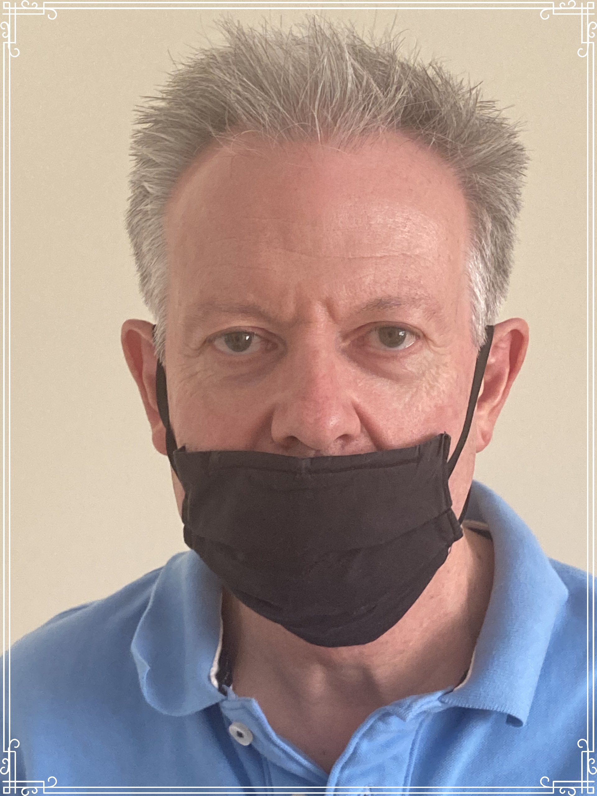 A person with graying hair wearing a blue polo shirt and a black face mask, captured in a head-and-shoulders portrait.