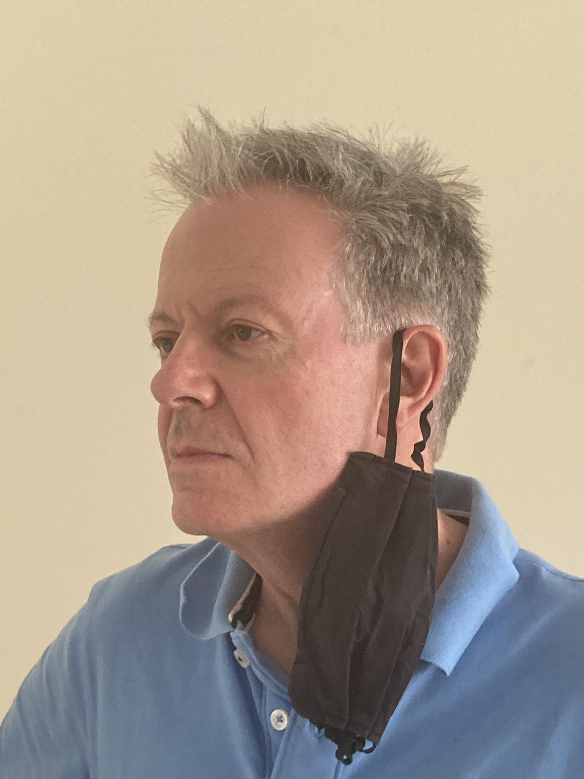 A person with short, light-colored hair wearing a blue polo shirt, with a black face mask hanging from one ear.