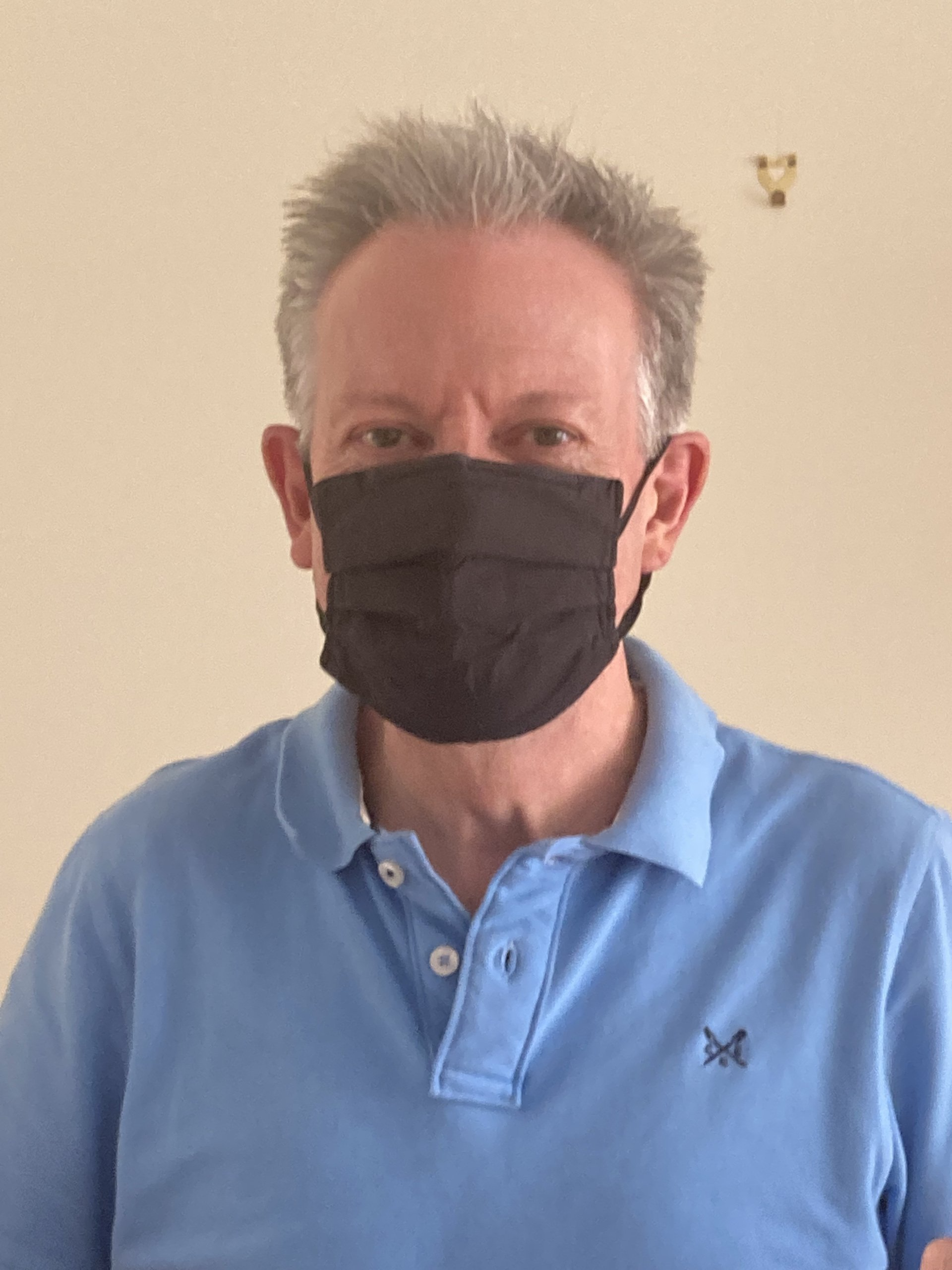 A person wearing a black face mask and a light blue polo shirt, standing against a plain, light-colored wall.