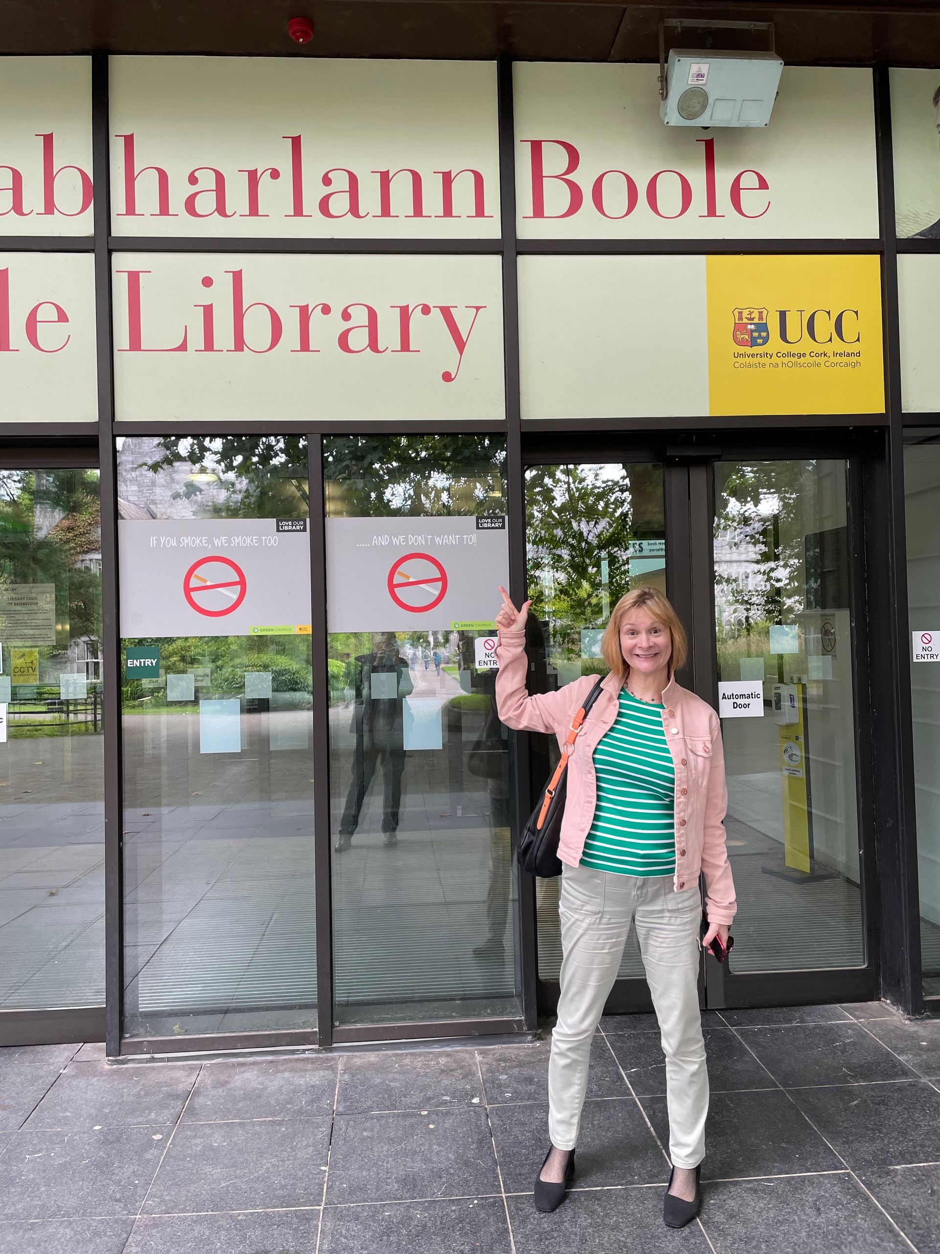 A smiling woman stands in front of the Boole Library at University College Cork, giving a peace sign.
