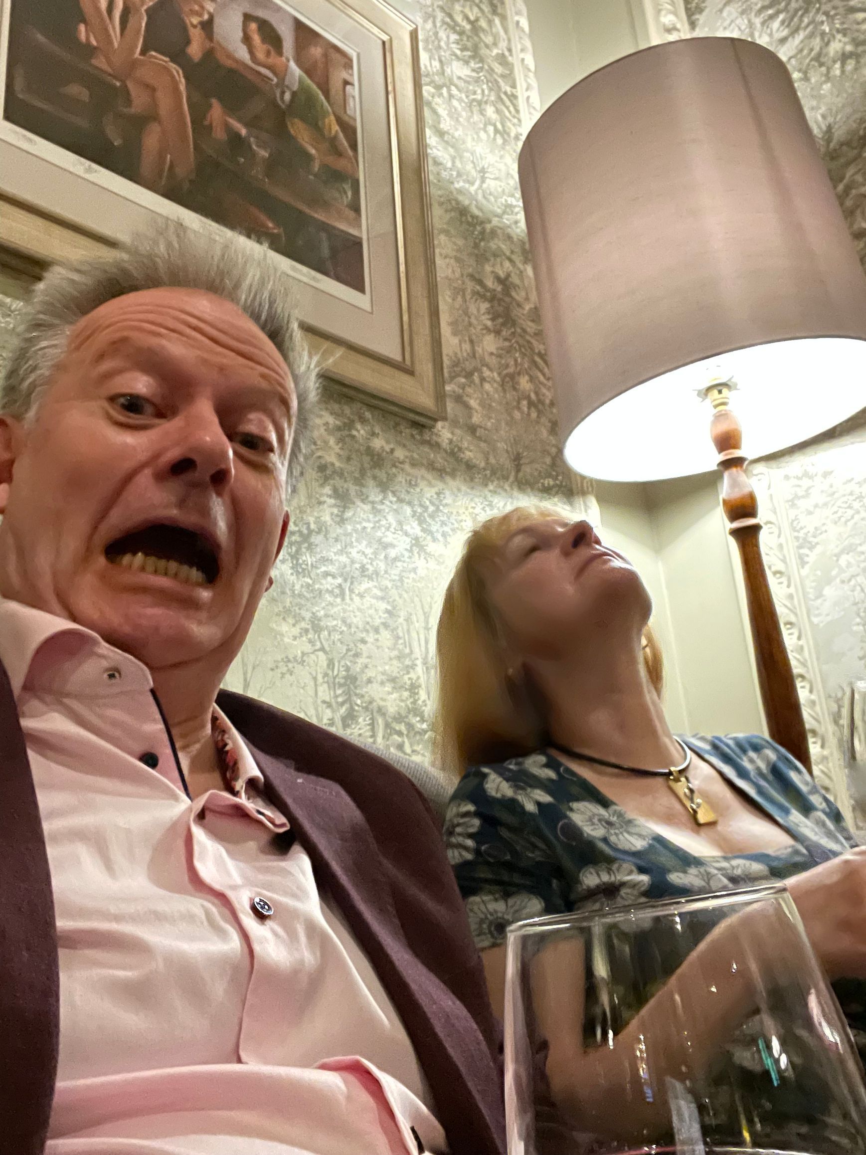 A man with a surprised expression poses for a selfie next to a woman leaning her head back, indoors under a floor lamp.