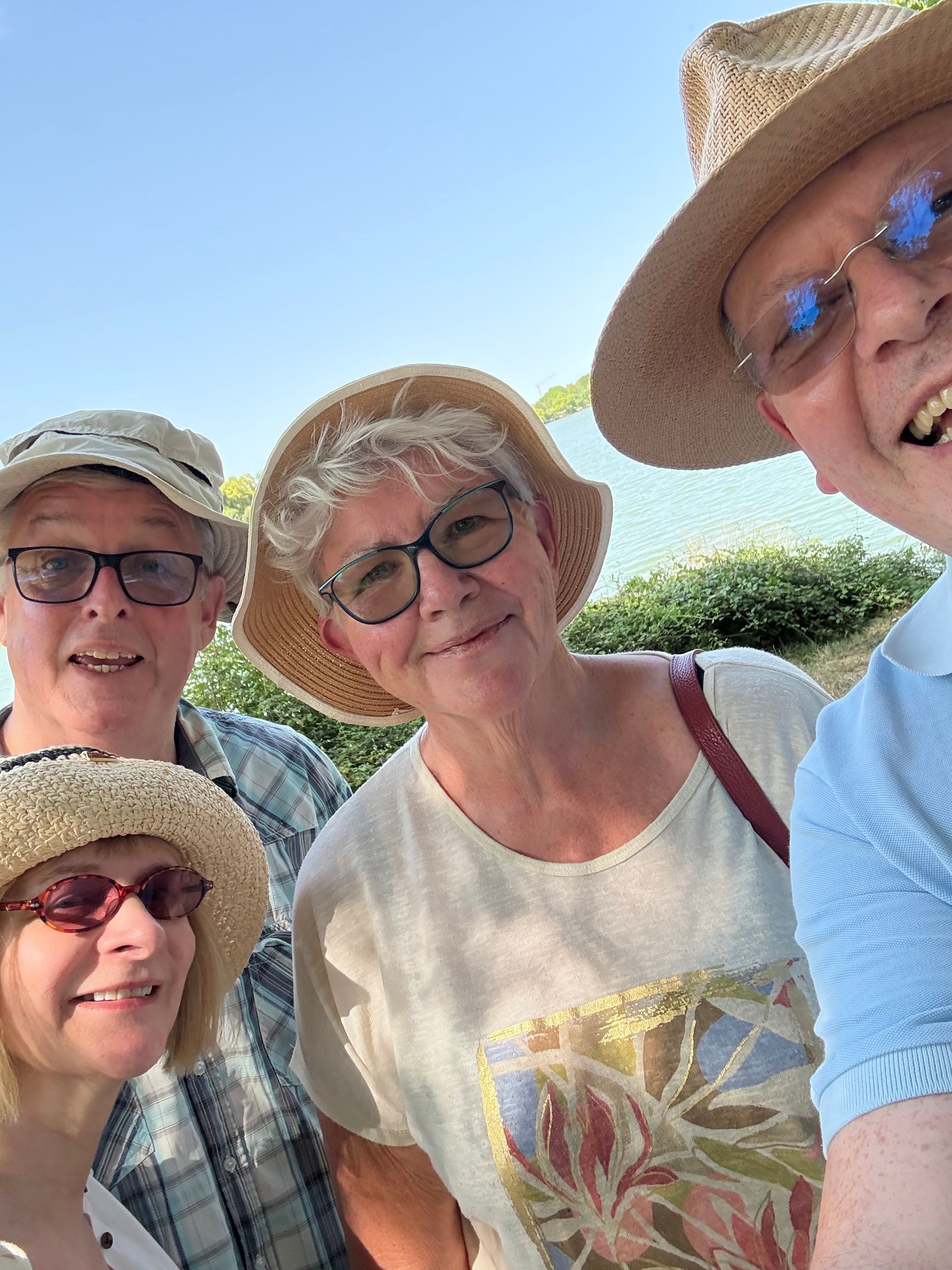 A low-angle, outdoor close-up of four people smiling at the camera, wearing sun hats and glasses under a clear blue sky.