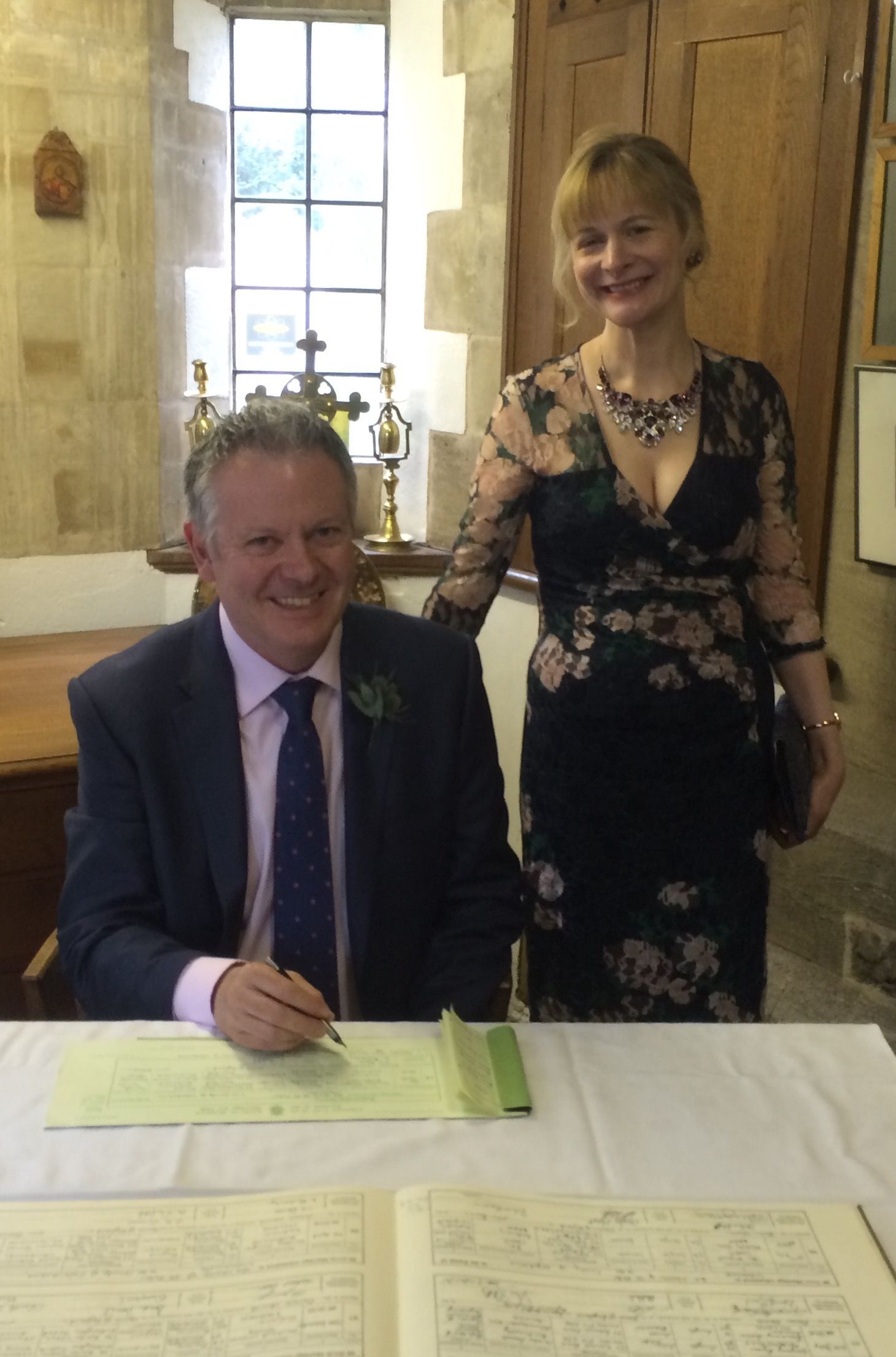 A smiling person in a suit signs a document at a table, while a person in a floral dress stands beside them in a church.