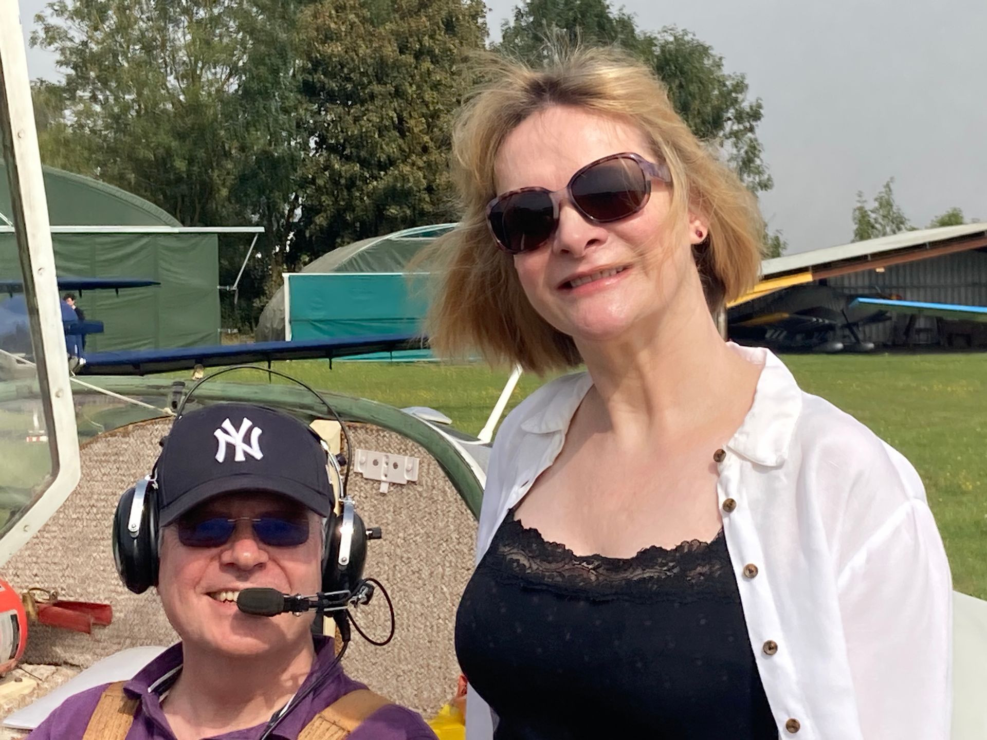 A person wearing a NY cap and headset sitting in a small aircraft next to a smiling person wearing sunglasses outdoors.