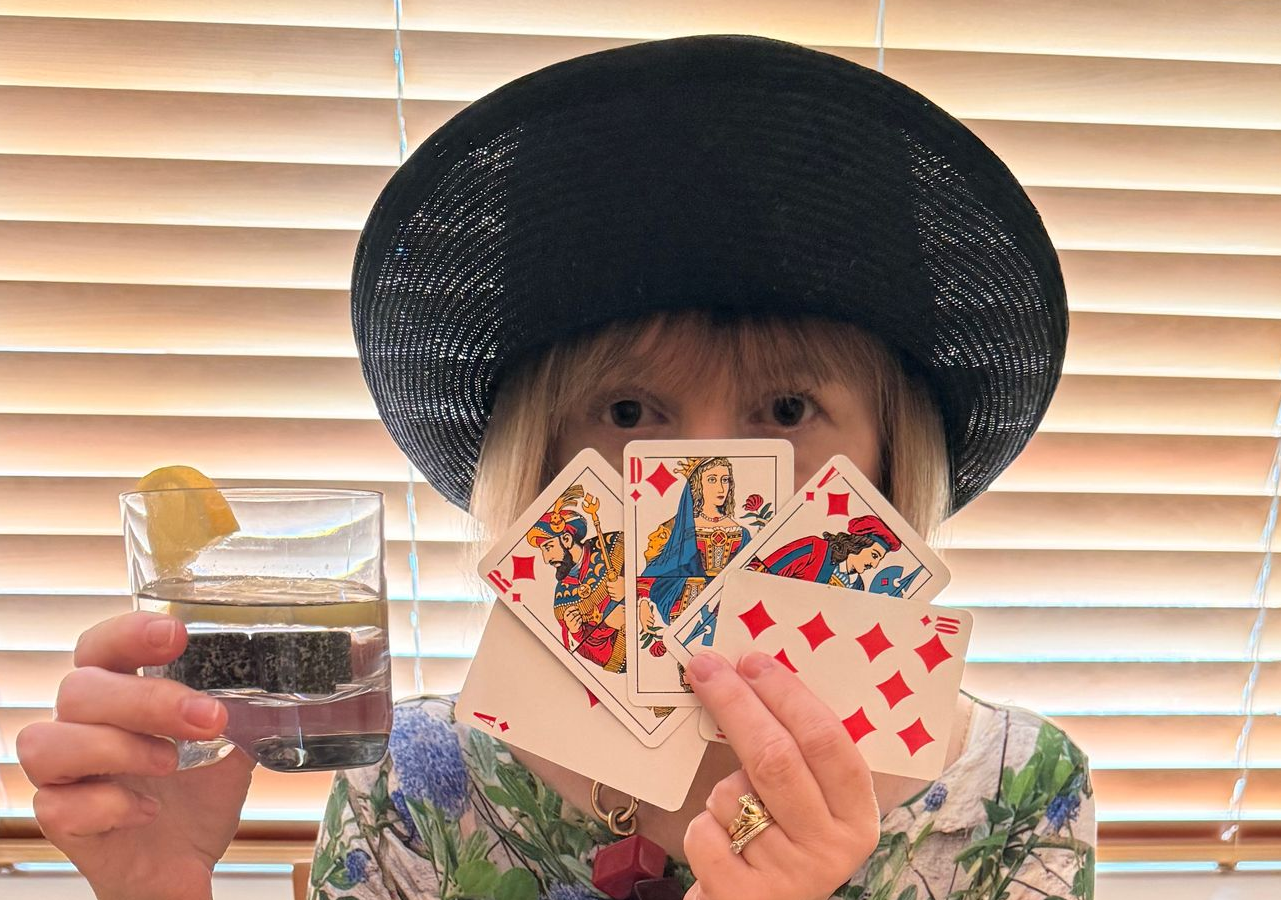 Woman in black hat and green-patterned Agnès B top, holding a hand of playing cards and a drink.