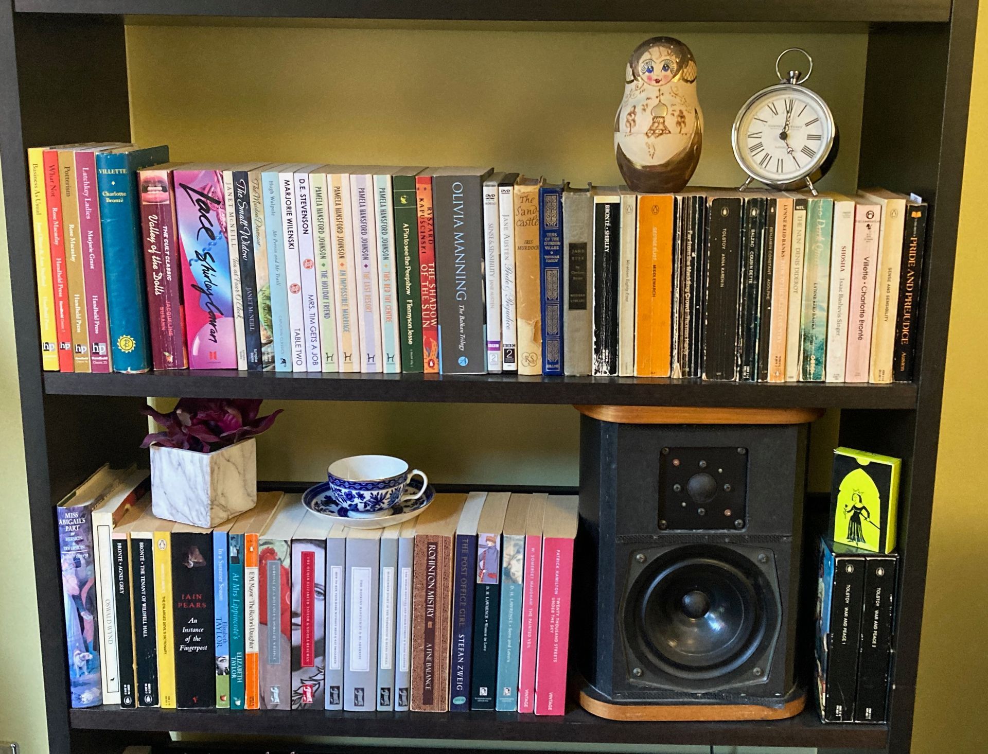 Two shelves of a bookcase filled with colorful books, a nesting doll, an analog clock, a small plant, and a speaker.