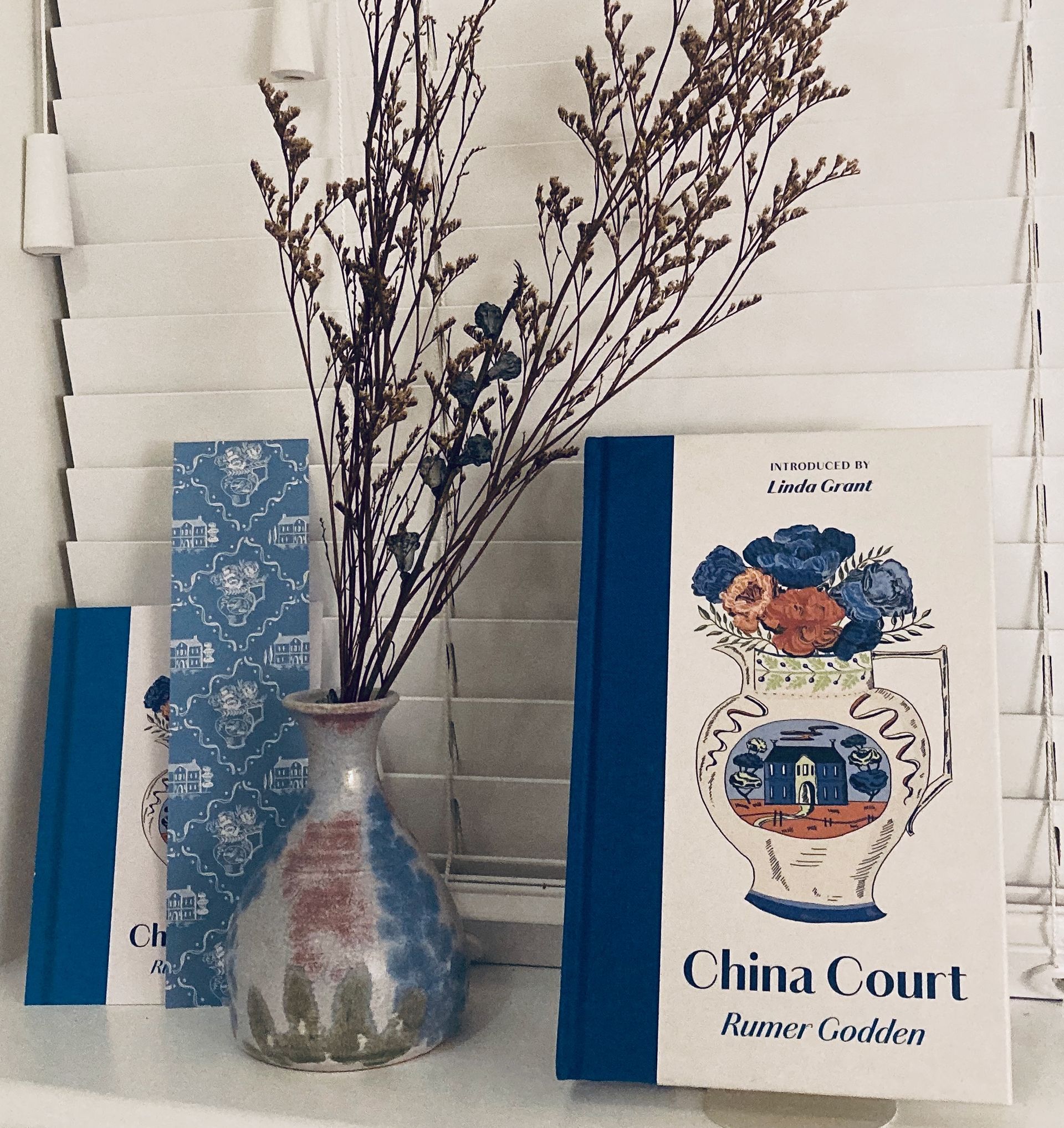 A vase of dried flowers stands between two copies of the book China Court by Rumer Godden against a white blind background.