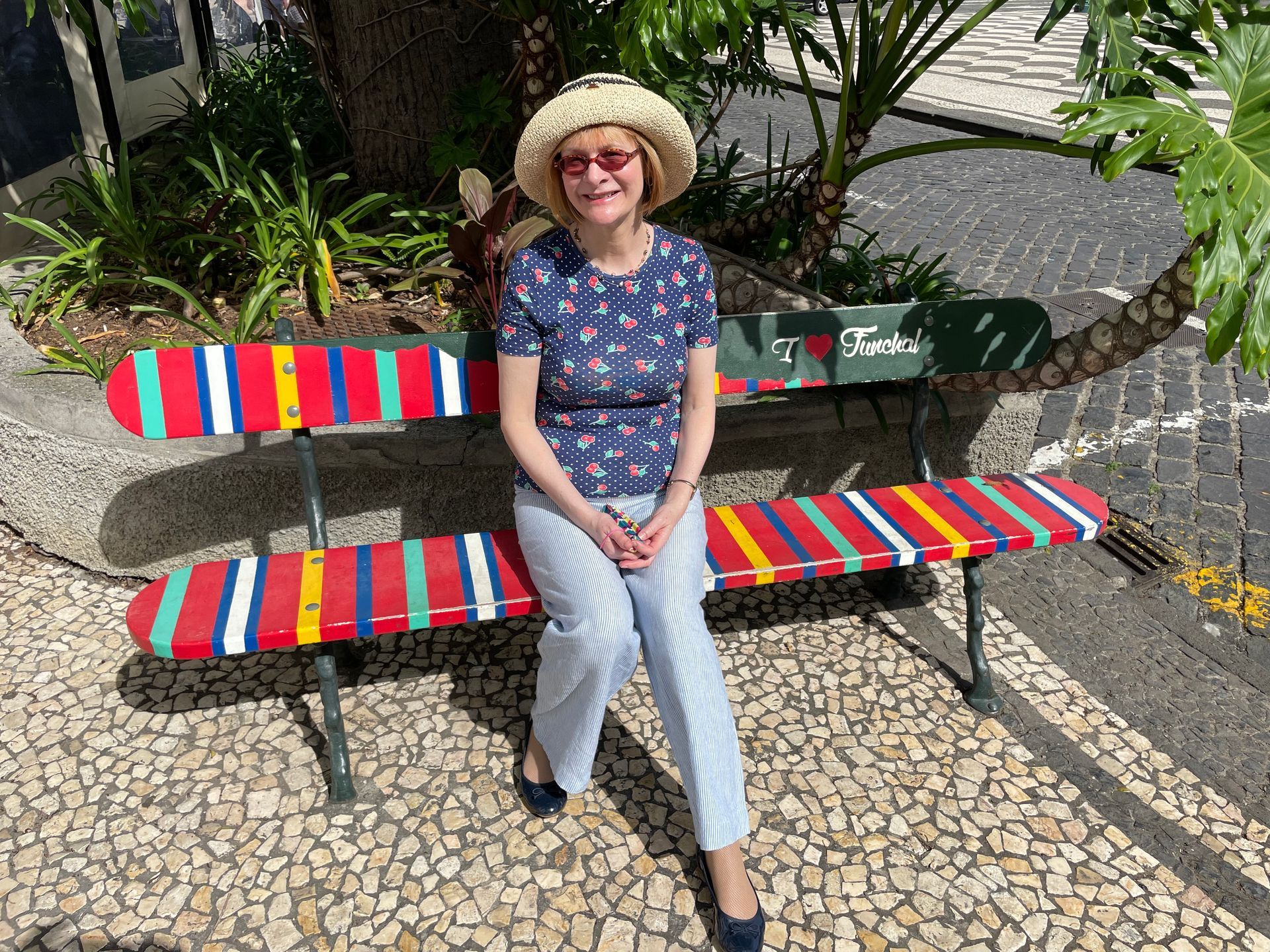 A person wearing a straw hat and floral top sits on a colorful, striped park bench on a stone-paved sidewalk.