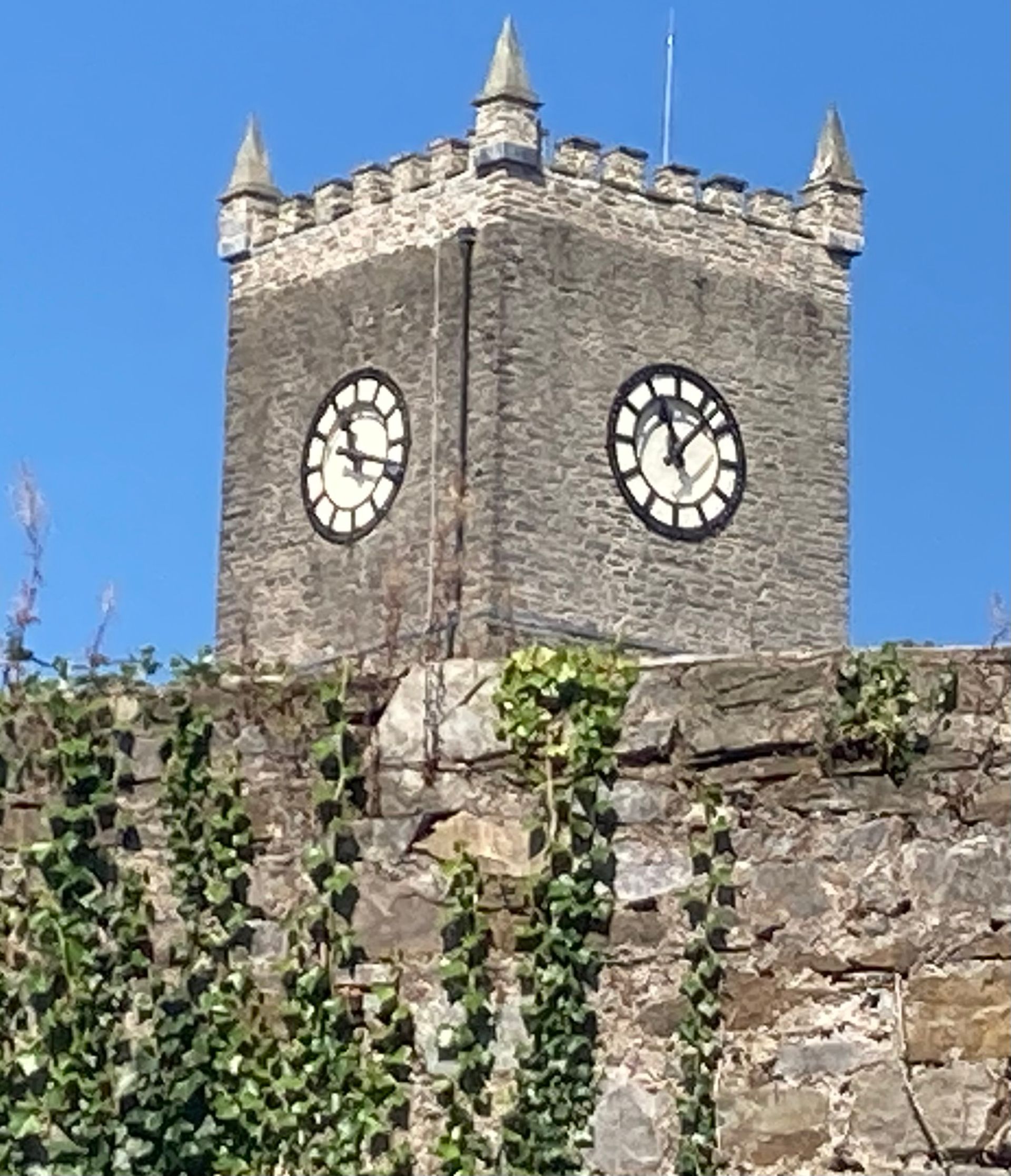 A stone clock tower with four turrets rises above a stone wall covered in climbing ivy against a clear blue sky.