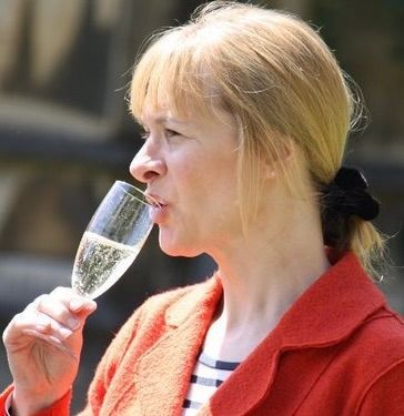 A person with blonde hair in a ponytail and a red blazer drinking from a flute glass of sparkling wine.