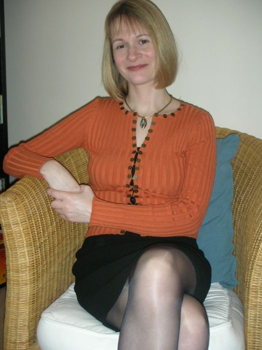 A person with short blonde hair wears an orange ribbed cardigan and a black skirt while sitting in a wicker armchair.