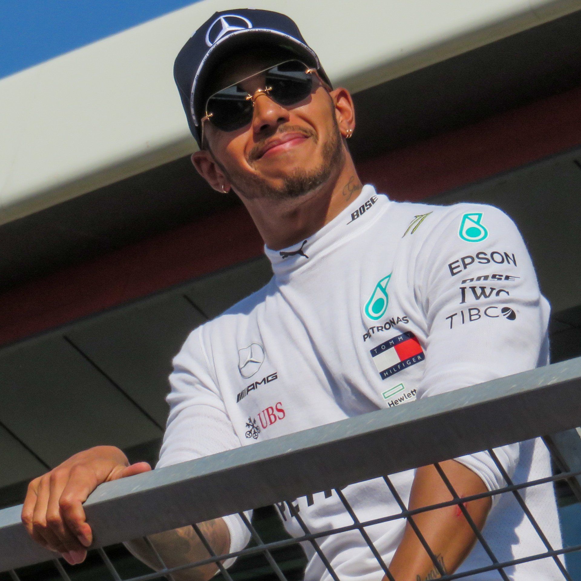 Lewis Hamilton wearing a white Mercedes racing team shirt, a team cap, and sunglasses, leaning against a metal railing.