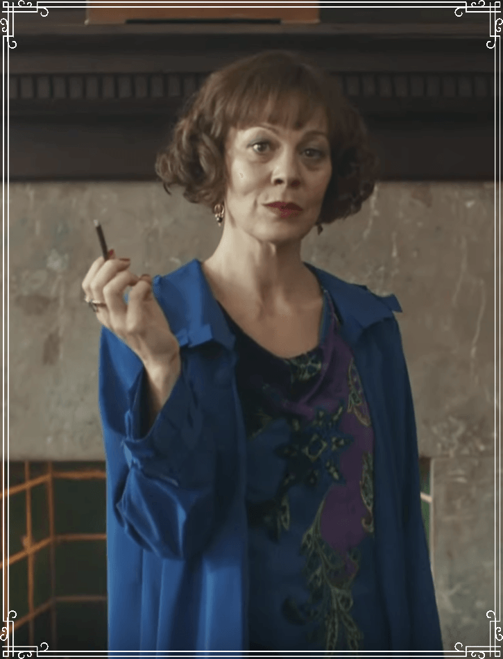 A person with short brown hair wears a blue jacket over a patterned top, holding a small object in front of a fireplace.