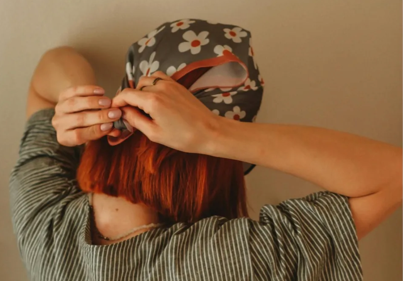 A person with reddish-brown hair ties a gray floral headscarf behind their head, wearing a gray striped shirt.