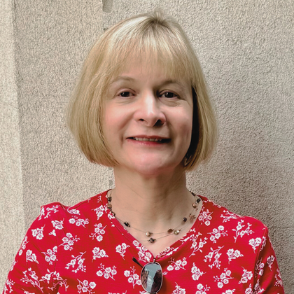 A person with light hair and a soft smile wearing a red floral print shirt and a beaded necklace against a textured wall.
