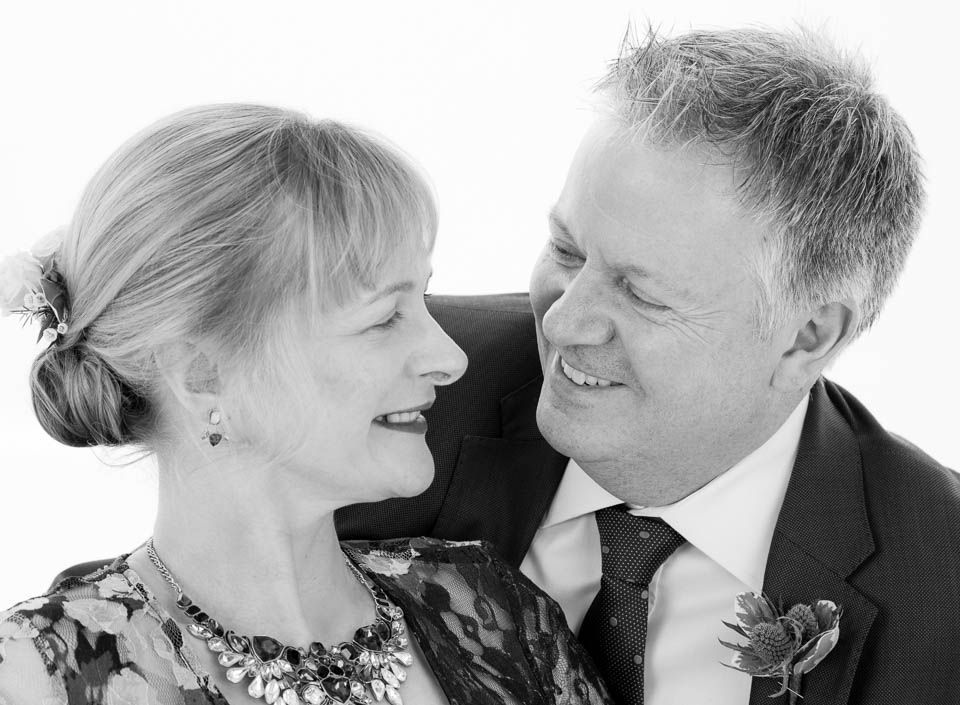 A couple in formal attire looks at each other with warm smiles in a black-and-white studio portrait.