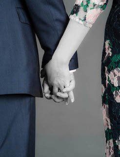 Two people in formal attire, a dark suit and a floral dress, holding hands against a neutral gray background.