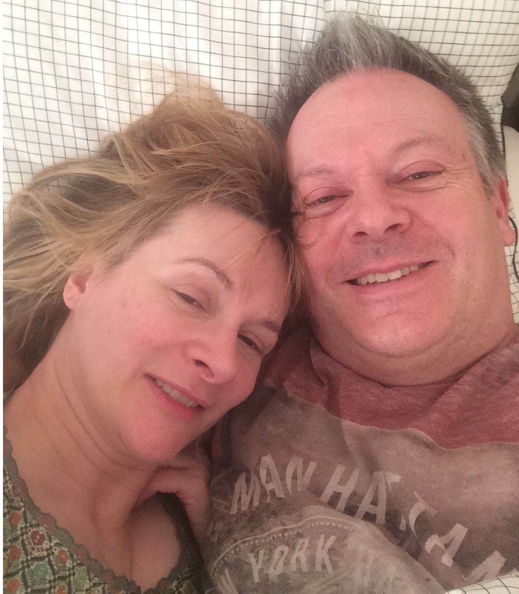 Two people lie together, smiling softly while looking at the camera against a patterned background.