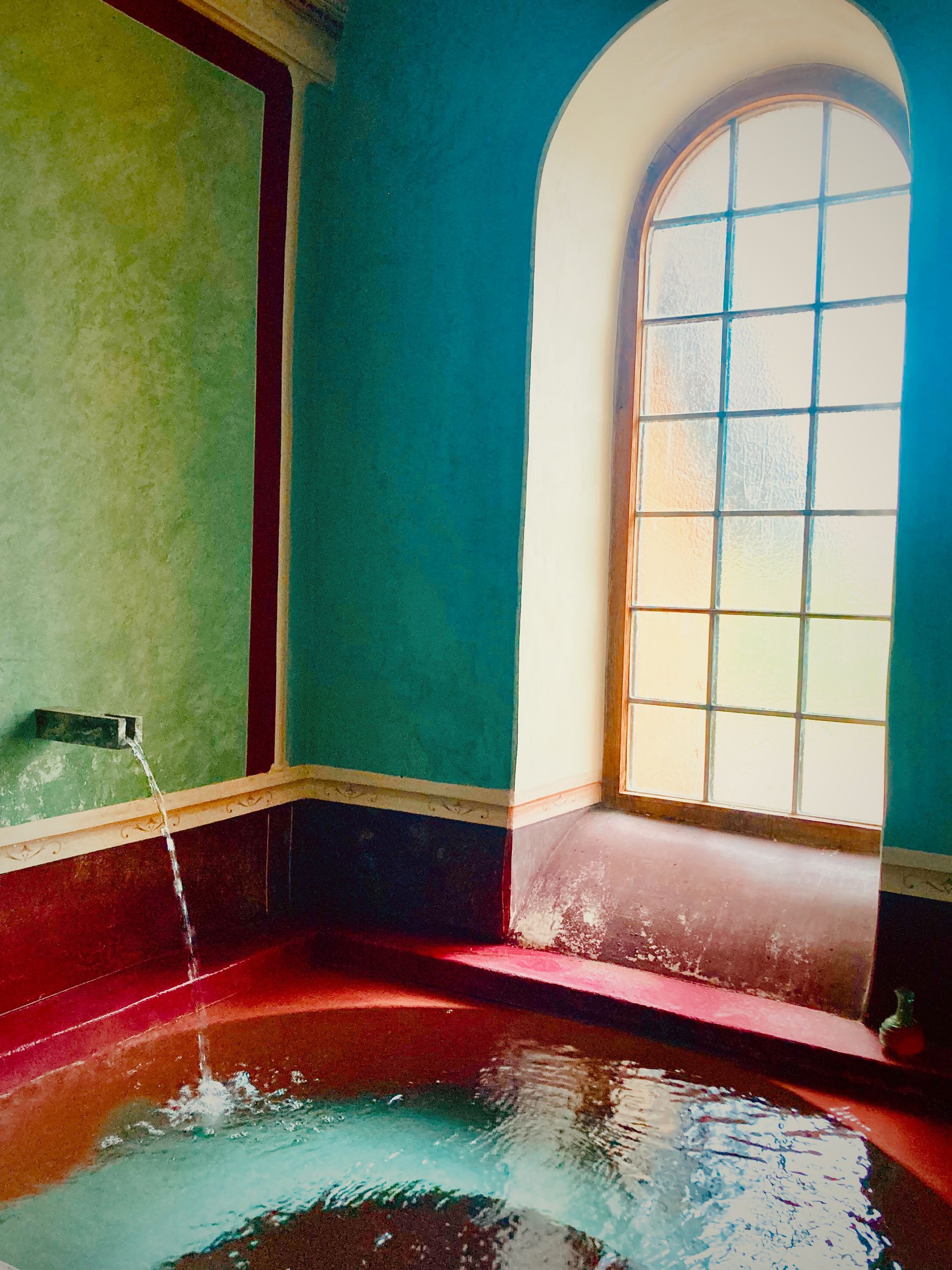 A red circular tub filled with water, featuring a wall-mounted faucet, inside a room with teal walls and an arched window.