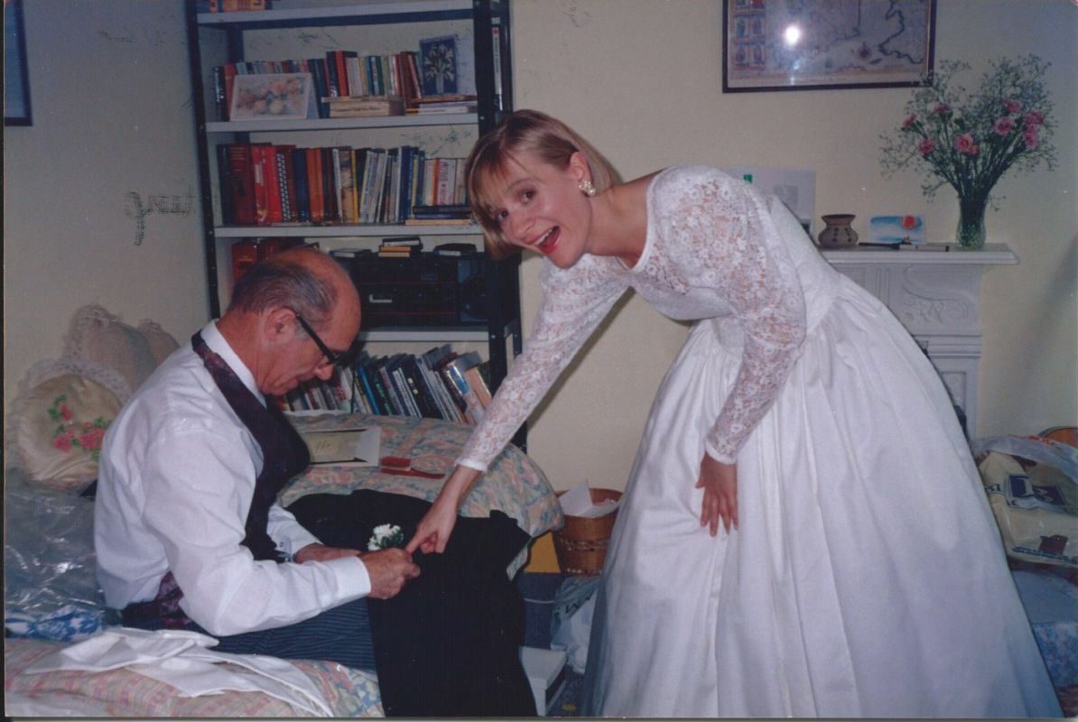 A person in a white wedding gown leans over to pin a boutonniere onto a seated person wearing a white shirt and vest.
