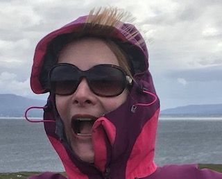 A person with a surprised expression wears a purple and pink rain hood and sunglasses against a coastal backdrop.