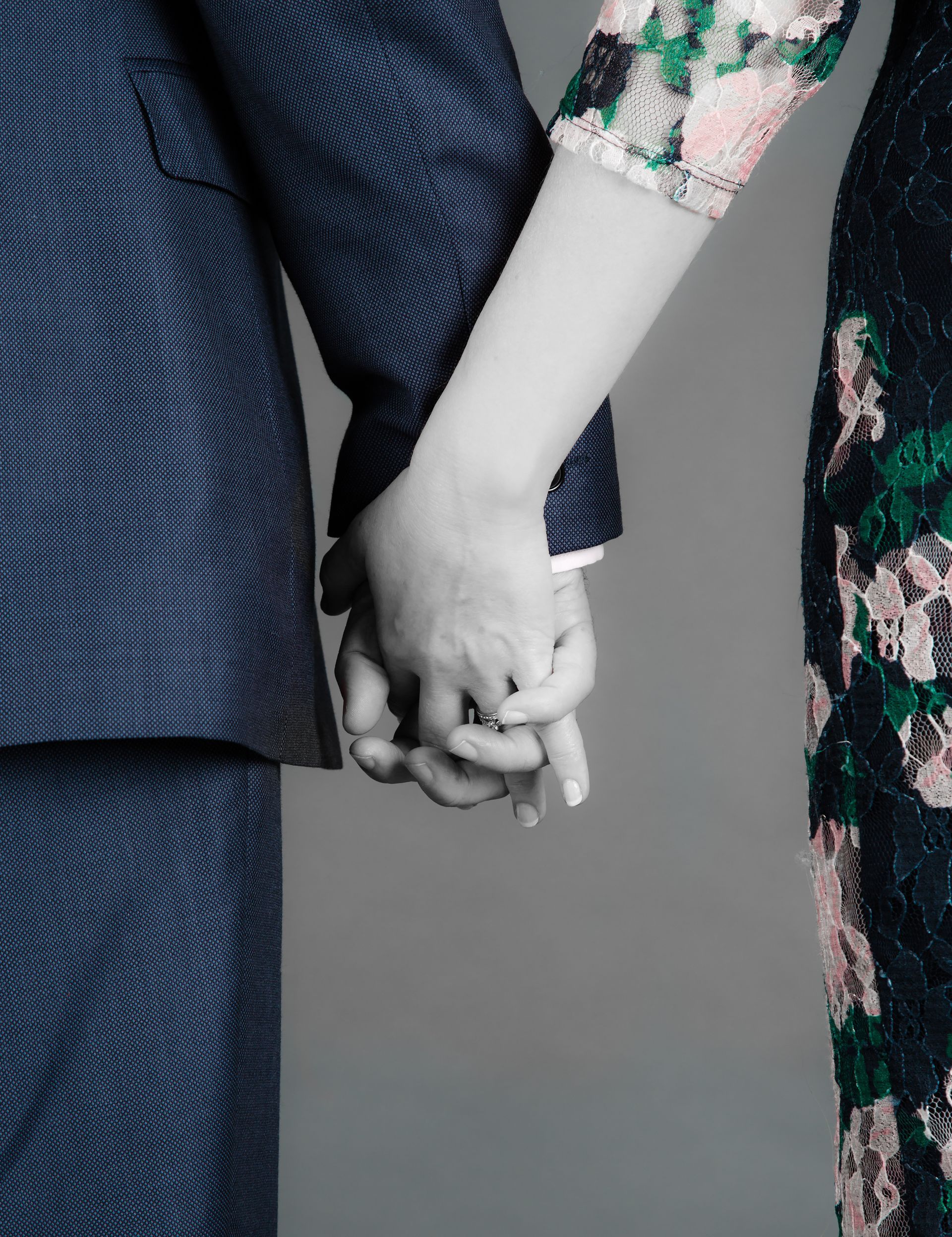Two people wearing formal attire, one in a dark patterned suit & one in a floral dress, hold hands against a gray background.