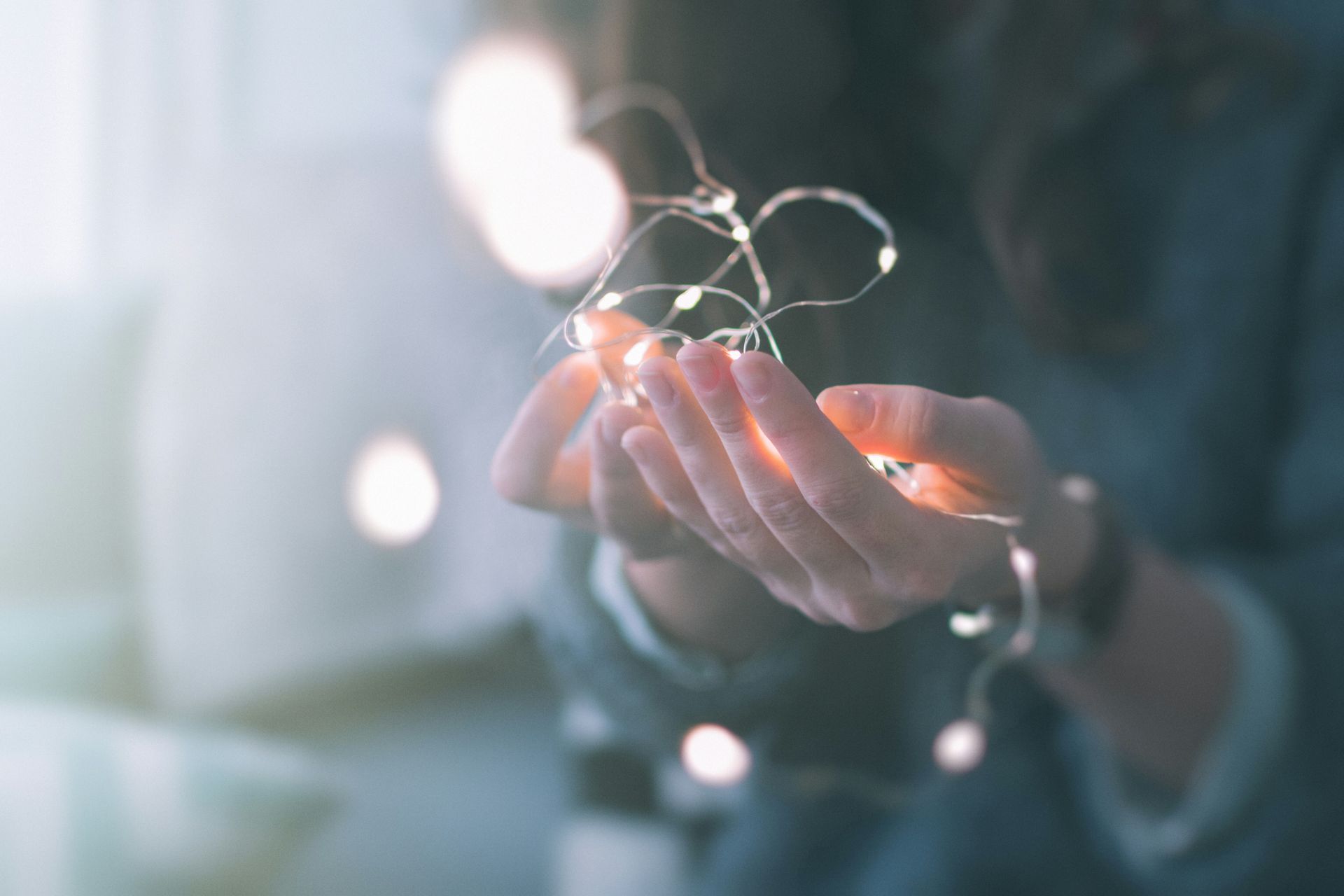 Cupped hands hold a strand of glowing fairy lights against a softly blurred, neutral background.