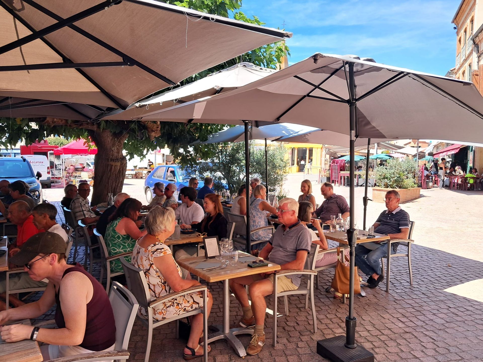 People dining at outdoor tables under large umbrellas on a sunny, paved town square.