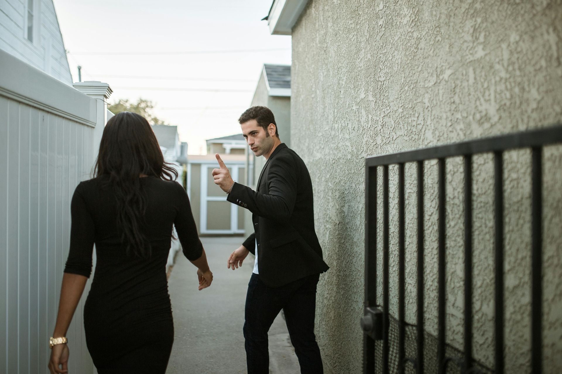 A person in a black outfit walks away while another person in a dark jacket points a finger toward them in an alleyway.