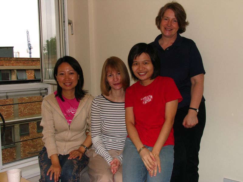 Four people pose together indoors against a plain wall, with a view of a city building through a window to the left.