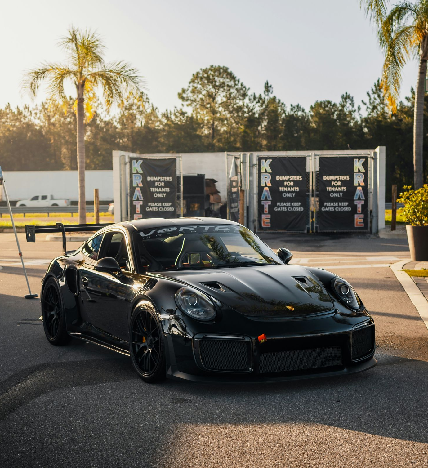 Black Porsche 911 GT2 RS parked near a container displaying a karting advertisement, palm trees in the background.
