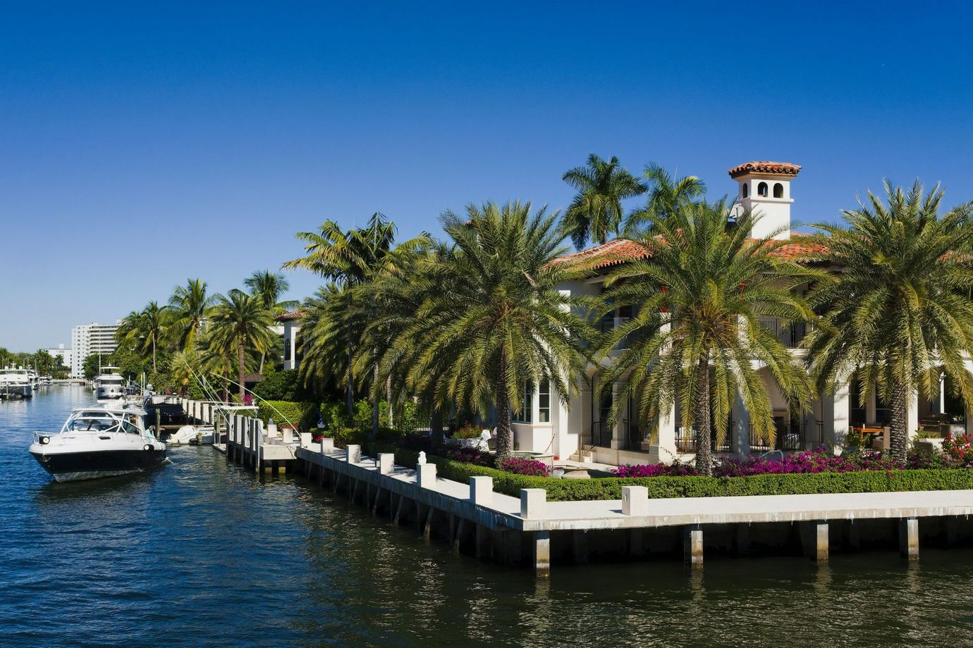 Waterfront mansion with palm trees, boat docked, blue sky.
