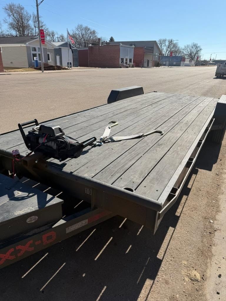 Flatbed trailer in a gravel lot with a black strap and equipment on top, buildings in the background