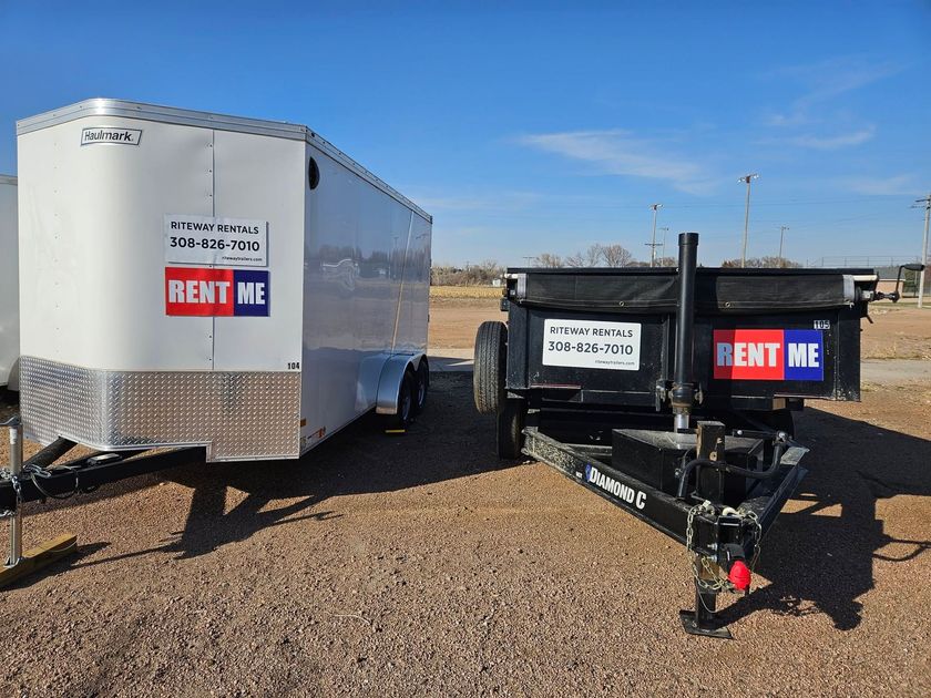 White enclosed trailer and black dump trailer with RENT ME signs on a gravel lot under blue sky