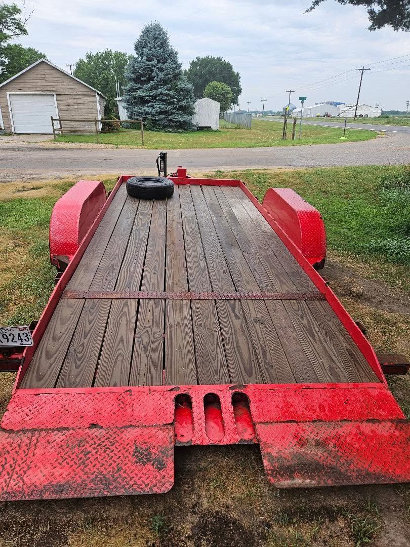 Red flatbed trailer with wooden deck and wheel arches parked on grass near a road.