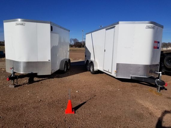 Two white enclosed trailers parked on a gravel lot with an orange cone in front of them.